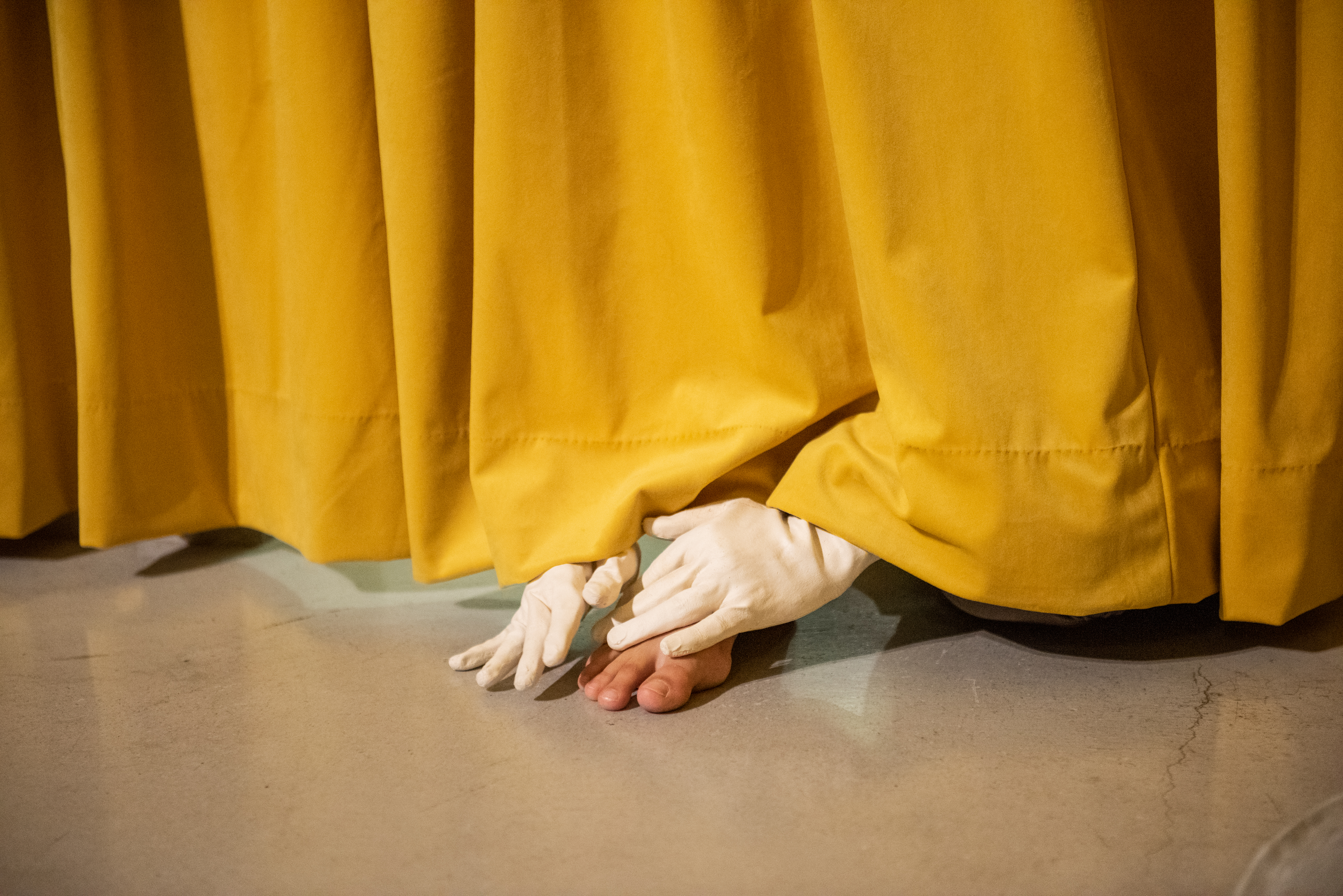 twee handen met latex handschoenen en een voet steken onder een geel gordijn uit